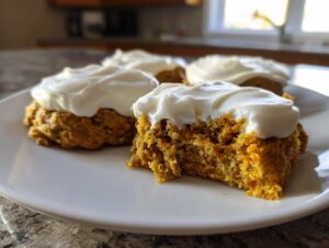 Close-up of soft carrot cake cookies topped with thick cream cheese frosting; one cookie is broken open showing texture.
