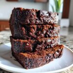 Four thick slices of moist chocolate banana bread, loaded with chocolate chips, stacked on a white plate.