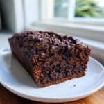 A freshly baked, dark chocolate zucchini bread loaf topped with melted chocolate chips on a white plate.
