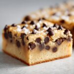 A close-up of a thick, square piece of salted caramel fudge loaded with chocolate chips and topped with sea salt.