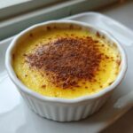 Close-up of a creamy yellow egg custard dessert topped with a dusting of brown spice, likely cinnamon, in a white ramekin.