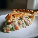 A close-up slice of chicken pot pie recipe showing flaky crust and creamy filling with shredded chicken, peas, and carrots.