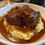 Two chunks of tender beef covered in rich brown gravy served over mashed potatoes, part of the slow cooker beef manhattan recipe.