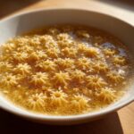A close-up shot of star-shaped pastina soup floating in golden broth within a white bowl.