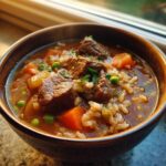 Close-up of a steaming bowl of beef barley soup, showing chunks of beef, carrots, peas, and barley.