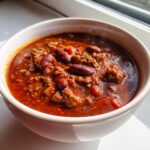 Close-up of a steaming white bowl filled with a rich, thick chili recipe featuring ground beef and kidney beans.