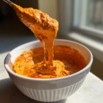 Close-up of thick, orange chili cheese dip being lifted from a white bowl with a spoon, showing its stretchy texture.