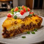 A hearty slice of taco pie showing layers of seasoned beef, cheesy crust, topped with sour cream, diced tomatoes, and green onions.