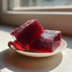 Two glistening, deep red cubes of homemade Yokan resting on a small white saucer, backlit by sunlight.