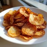 A close-up of a mound of crispy, seasoned air fryer potato chips piled high on a white plate.