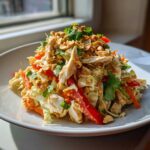 A vibrant mound of thai chicken salad featuring shredded chicken, cabbage, red peppers, and topped with crushed peanuts and cilantro.