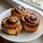 Three freshly baked banana cinnamon rolls twist covered in rich caramel glaze, sitting on a white plate.