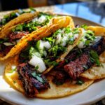 Close-up of three freshly made Carne Asada Tacos topped with diced white onion and fresh cilantro on a white plate.