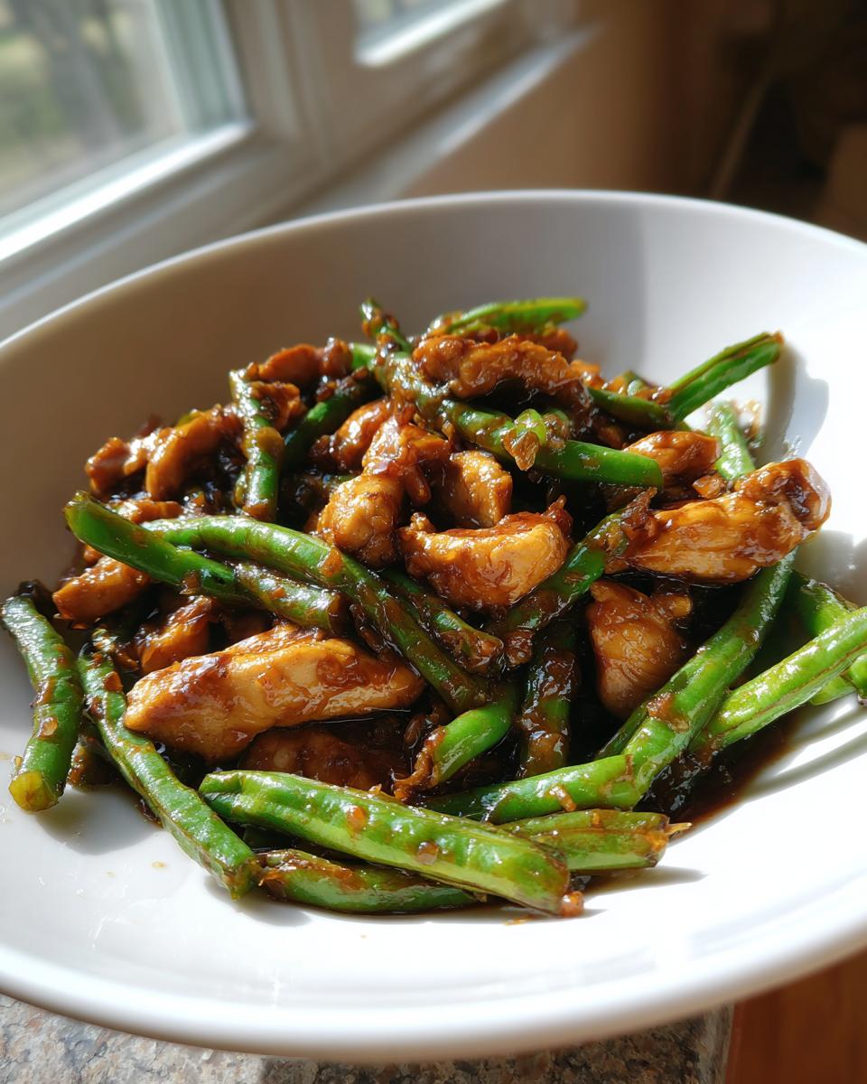 A close-up view of glossy, saucy chicken and green bean stir fry served in a white bowl near a window.