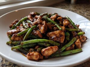 Close-up of a white bowl filled with glossy chicken and green bean stir fry coated in dark sauce.