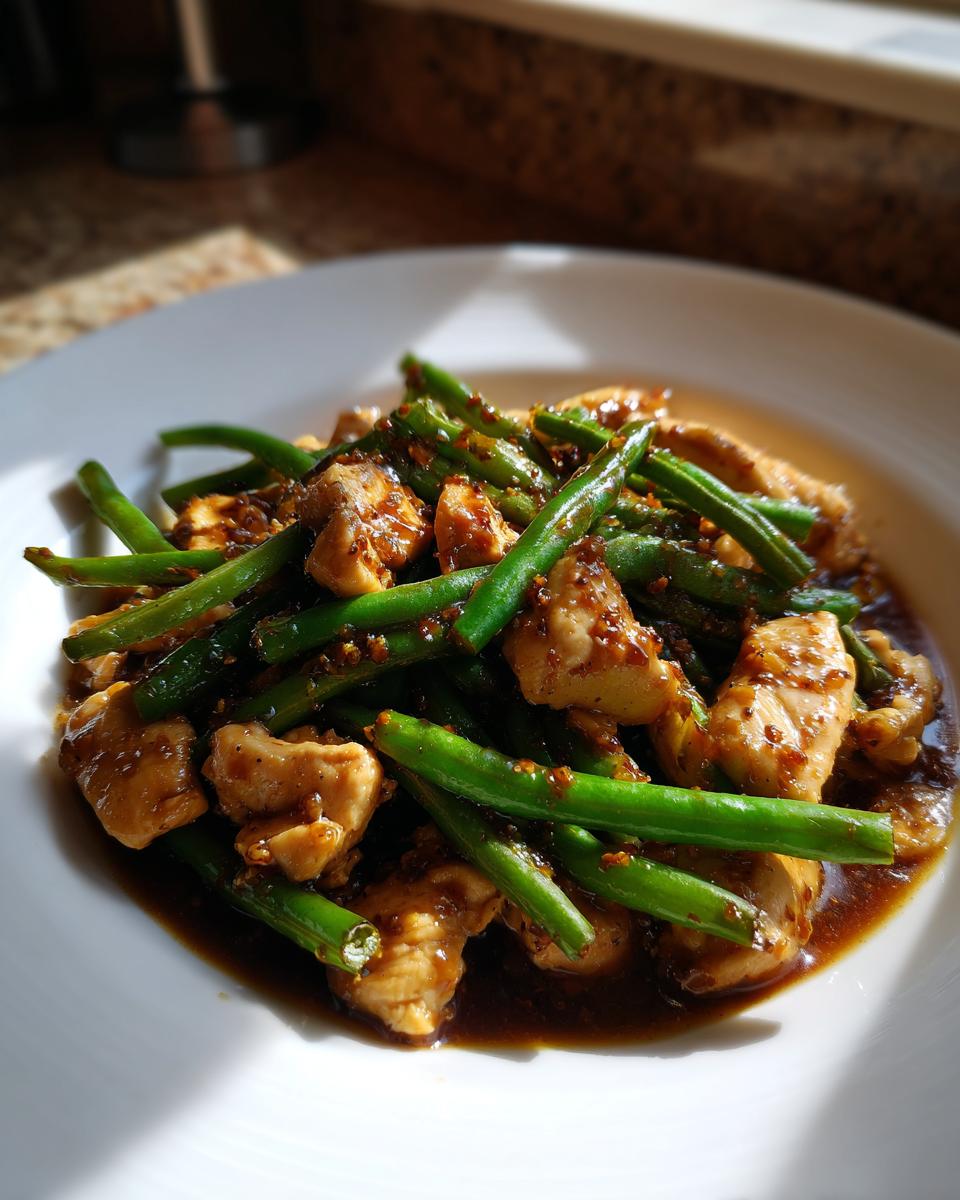 A close-up of chicken and green bean stir fry coated in a rich brown sauce, served on a white plate.