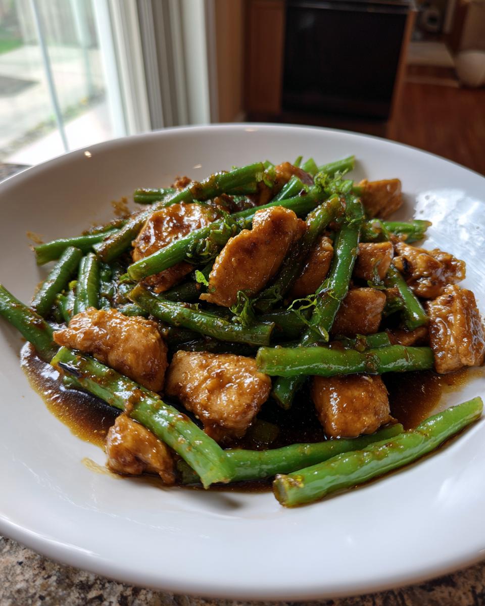 Close-up of a white bowl filled with glossy chicken and green bean stir fry coated in a savory brown sauce.