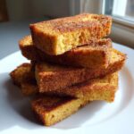 A tempting stack of golden brown cinnamon french toast sticks heavily coated in cinnamon sugar, resting on a white plate.