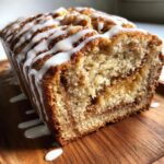 A close-up of a loaf of cinnamon roll banana bread, sliced to show the swirl, drizzled heavily with white icing.