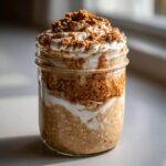 Close-up of layered cinnamon roll overnight oats topped with cream and brown sugar crumble in a glass jar.