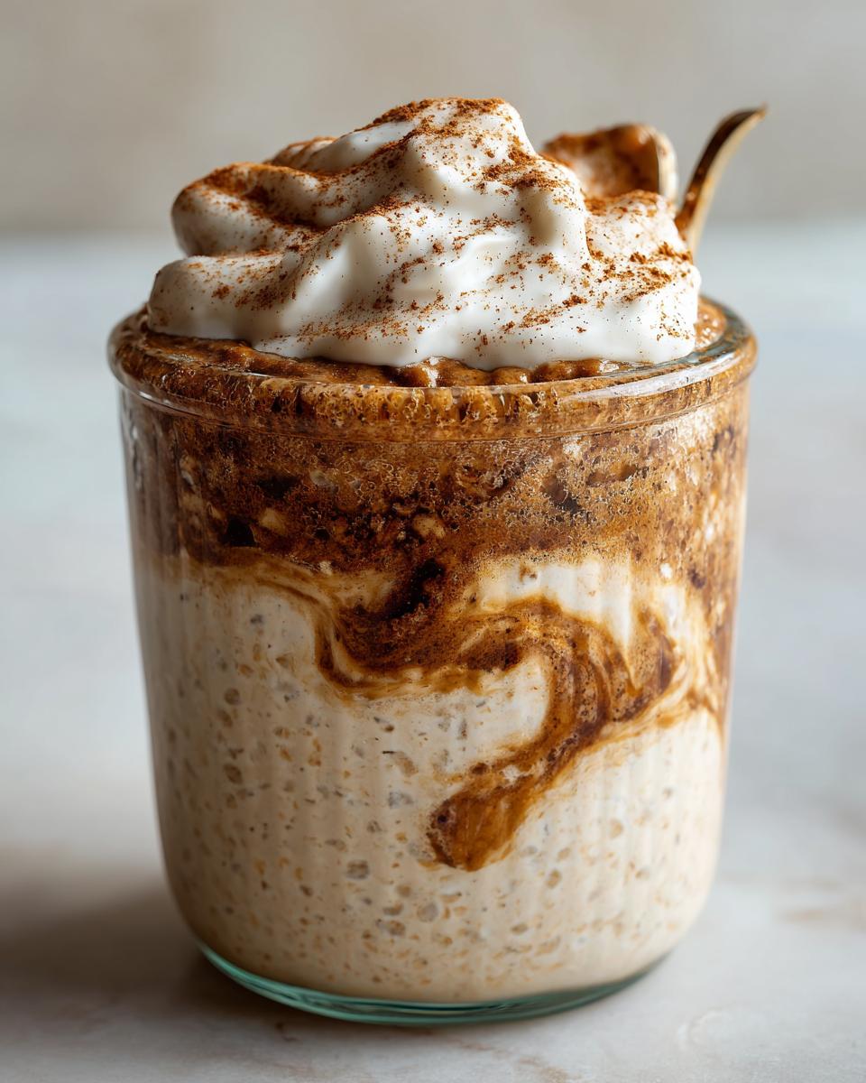A glass jar filled with creamy cinnamon roll overnight oats, swirled with cinnamon mixture and topped with whipped cream and extra spice.
