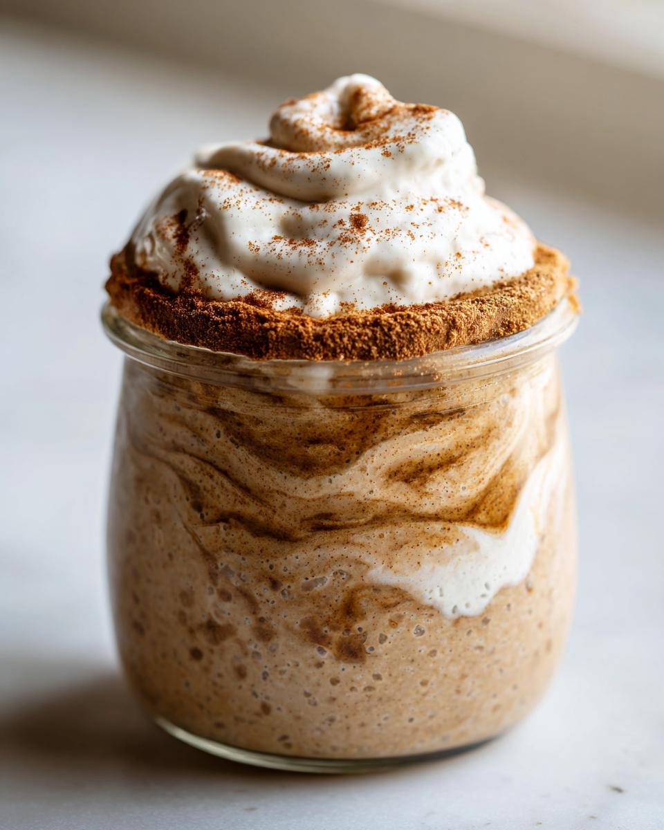 A glass jar filled with swirled cinnamon roll overnight oats, topped with a generous swirl of white cream and sprinkled with cinnamon.