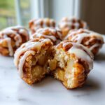 A close-up of one apple fritter bite cut in half, showing soft interior and chunks of apple, drizzled with white glaze. More apple fritter bites are in the background.