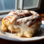 A close-up of a single, fluffy cinnamon sugar rolls covered in thick white icing, sitting on a white plate.