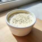 A small white ramekin filled with rich, creamy alfredo sauce seasoned heavily with cracked black pepper.