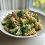 A close-up of creamy Broccoli Pasta Salad featuring rotini pasta, bright green broccoli florets, and crumbled bacon served in a white bowl.