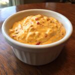 Close-up of a creamy, orange-colored cheese dip with visible red pepper chunks served in a white ramekin.