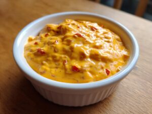A close-up of a creamy, bright orange cheese dip with visible red pepper pieces served in a small white ramekin.