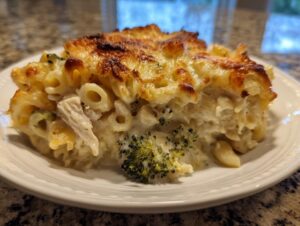 A close-up of a serving of creamy chicken alfredo pasta bake featuring a golden, bubbly cheese crust and visible pieces of chicken and broccoli.