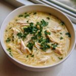 A close-up of a bowl of creamy chicken orzo soup, featuring shredded chicken and topped generously with fresh chopped parsley.