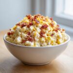 A creamy, heaped bowl of crack corn salad topped generously with shredded cheese and crispy bacon bits.