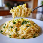 A spoonful of creamy lemon orzo pasta being lifted above a white bowl, garnished with parsley and zest.