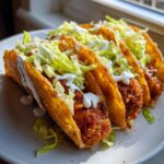Close-up of three golden, crispy potato tacos filled with seasoned potato and topped with shredded lettuce and a drizzle of white sauce.