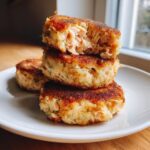 Close-up of three crispy, golden-brown salmon cakes stacked on a white plate, showing the flaky interior of the top salmon cake.