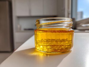 A small glass jar filled with golden Dandelion Honey, sitting on a bright white counter in sunlight.