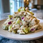 A close-up mound of creamy dill pickle chicken salad featuring shredded chicken, chopped pickles, and red onion on a white plate.