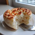 Close-up of a freshly cooked english muffin recipe with a bite taken out, showing its airy, porous interior.