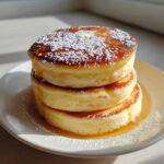 A tall stack of three thick, fluffy pancakes from scratch, drizzled with syrup and topped with powdered sugar and butter.