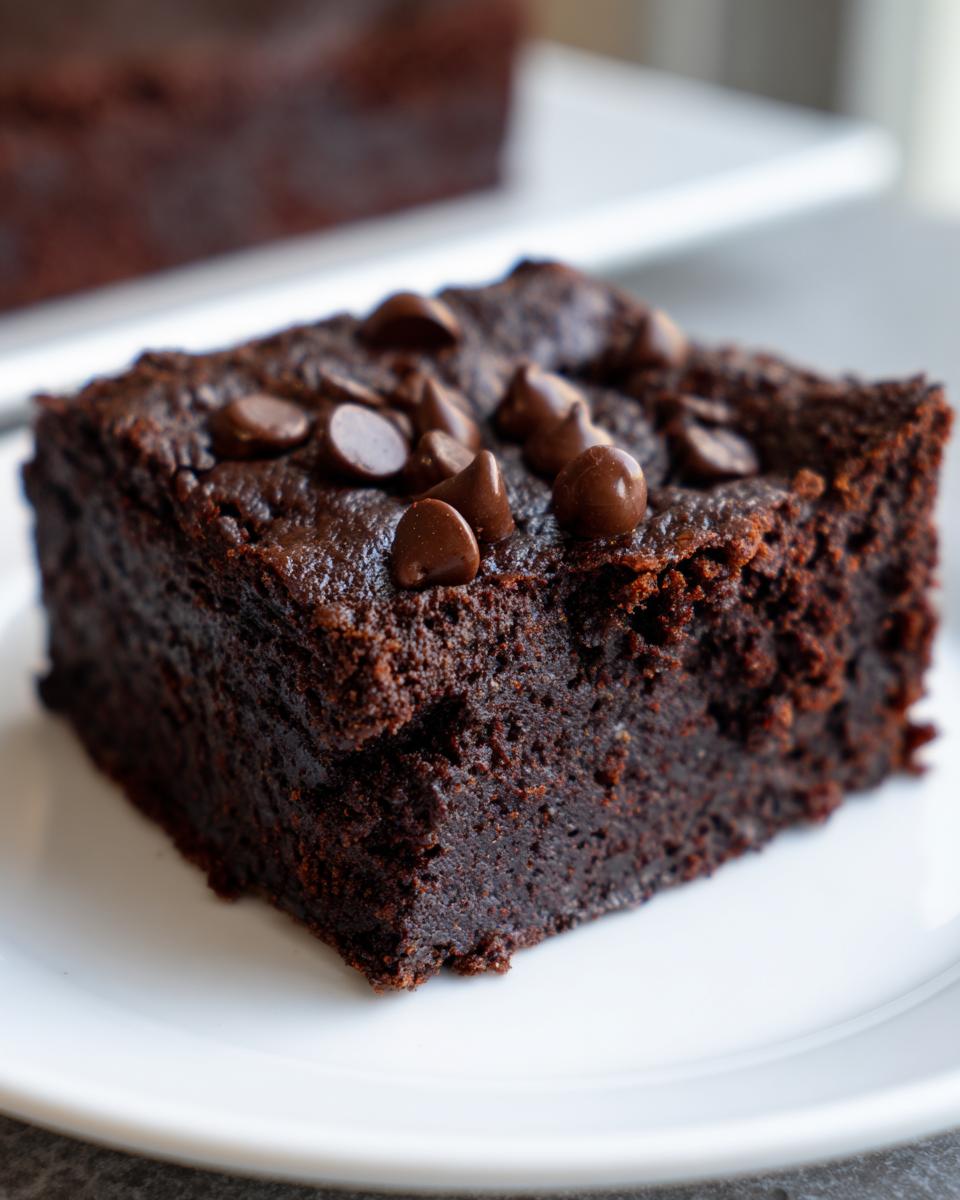 Close-up of a fudgy, dark chocolate date brownies square topped with chocolate chips on a white plate.