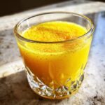 Close-up of a vibrant yellow Ginger Lemon Turmeric Shot topped with spice powder in a decorative glass.