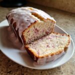 A loaf of glazed strawberry bread, partially sliced, showing the moist interior with visible strawberry pieces.