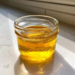 A small glass jar filled with bright, golden Dandelion Honey sitting on a white countertop in sunlight.