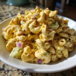 Close-up of a white bowl filled with creamy, yellow Guy Fieri's Iconic Mac Salad featuring elbow macaroni and red onion.
