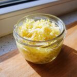 A small glass jar filled with bright yellow, shredded fermented cabbage sitting on a wooden cutting board.
