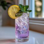 A tall glass filled with a bubbly, purple Lavender Mocktail, ice, garnished with a lemon wheel and fresh mint sprig.