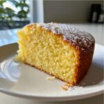 Close-up of a moist slice of bright yellow Lemon Ricotta Cake dusted with powdered sugar on a white plate.